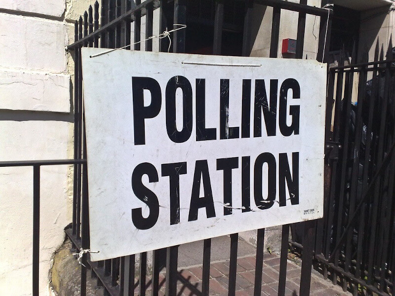a white board with large black letters POLLING STATION tied to black metal spikey fence