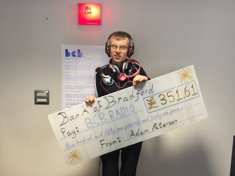 a man with many headphones draped over him, holding a representation of a cheque